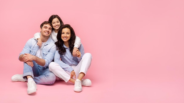 Pais e filha sentados em fundo rosa posando juntos e menina sorridente abraçando o pai