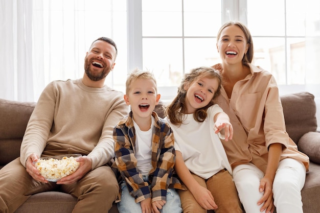 Pais e filhos da família encantados comendo pipoca e rindo enquanto estão sentados no sofá e assistindo filme engraçado em casa