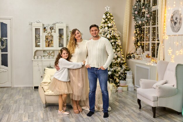 Pais felizes com a filha pequena em pé na sala de luz de ano novo decorado em casa no fundo da árvore de Natal. Família, conceito de férias