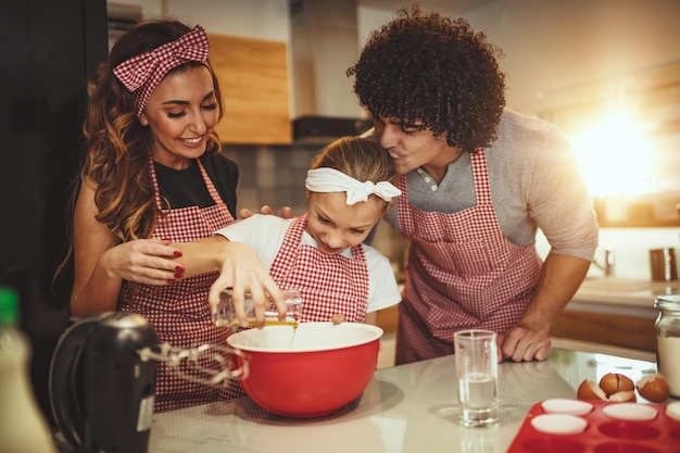 Pais felizes e sua filha estão preparando biscoitos juntos na cozinha. A menina ajuda seus pais derramando o óleo no prato com massa.