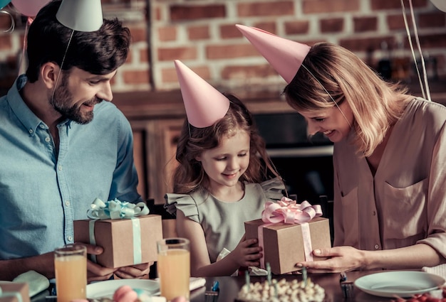 Pais felizes estão dando presentes para sua filha enquanto estão sentados à mesa na cozinha decorada durante a festa de aniversário