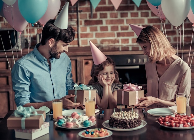 Pais felizes estão dando presentes para sua filha enquanto estão sentados à mesa na cozinha decorada durante a festa de aniversário