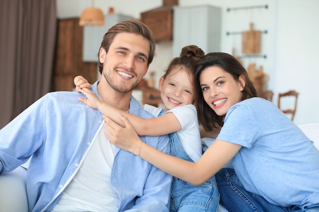 Pais jovens amigáveis positivos com a filha sorridente, sentados no sofá juntos, enquanto relaxam em casa no fim de semana.