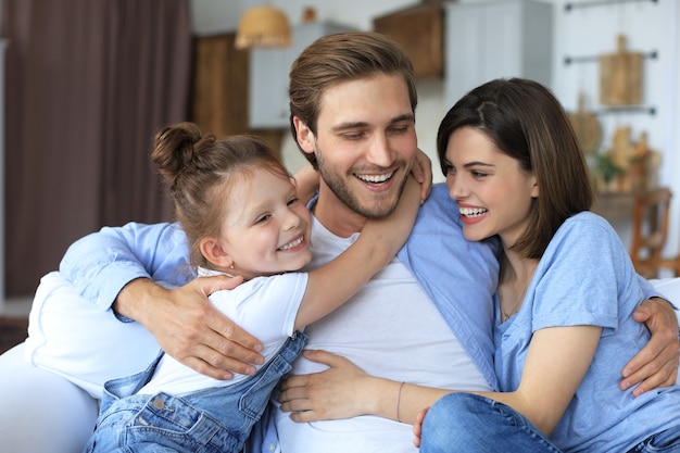 Pais jovens amigáveis positivos com a filha sorridente, sentados no sofá juntos, enquanto relaxam em casa no fim de semana.