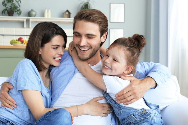 Pais jovens amigáveis positivos com a filha sorridente, sentados no sofá juntos, enquanto relaxam em casa no fim de semana.