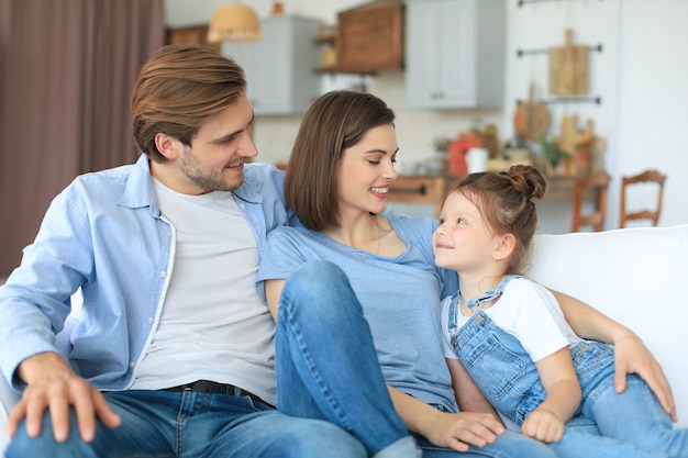 Pais jovens amigáveis positivos com a filha sorridente, sentados no sofá juntos, enquanto relaxam em casa no fim de semana.