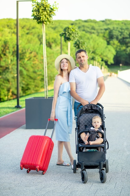 Pais jovens e felizes, mamãe e papai com uma mala vermelha e um bebê em um carrinho estão saindo de férias no verão, sorrindo