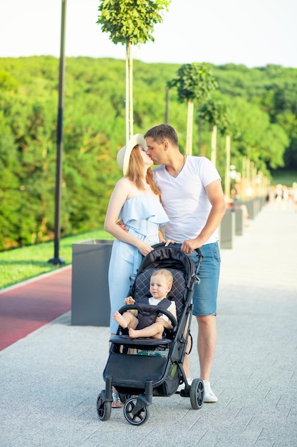 Pais jovens e felizes, mamãe e papai estão andando com um bebê em um carrinho no parque com um chapéu no verão no sol poente e sorrindo se divertindo