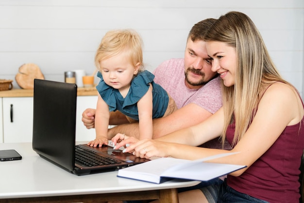 Pais jovens e uma criança pequena sentam em uma mesa sorrindo e olham para uma foto de alta qualidade de laptop