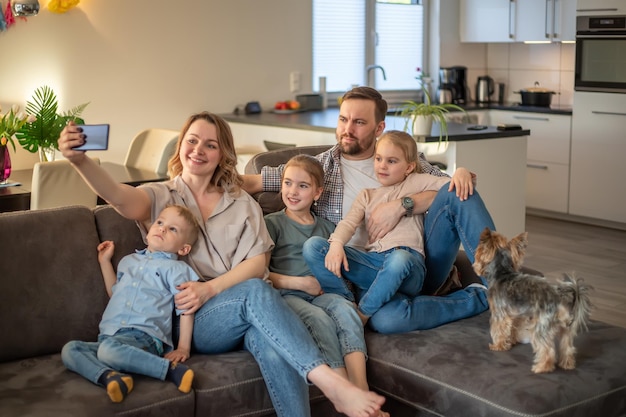 Pais jovens felizes sentam-se no sofá com crianças pequenas se divertem junto com a família caucasiana sorridente do smartphone assistem a vídeos engraçados no celular tirando uma selfie ou conversando em uma videochamada