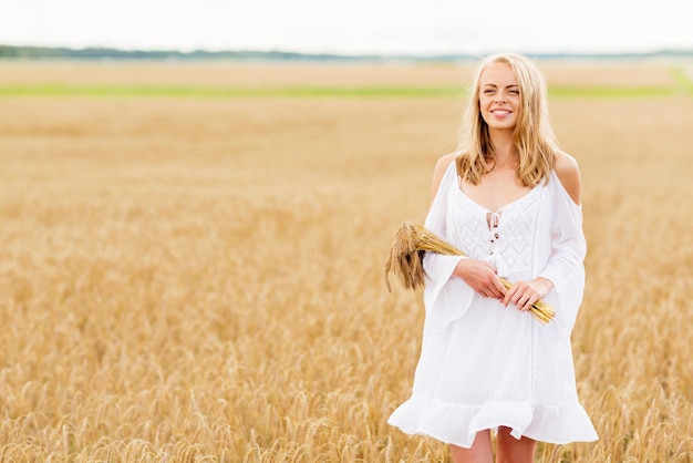 país, naturaleza, vacaciones de verano, vacaciones y concepto de la gente - joven sonriente vestida de blanco con espiguillas caminando por el campo de cereales