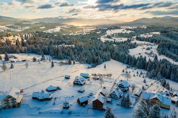 Paisagem aérea do inverno com pequenas casas de aldeia entre floresta coberta de neve em montanhas frias à noite.