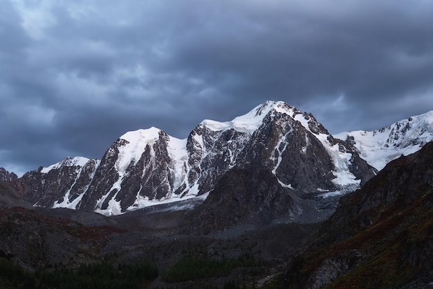 Paisagem alpina atmosférica escura com topo de montanha coberto de neve sob o céu noturno Paisagem incrível com belo pico pontiagudo com neve e parede de alta montanha nevada com nuvens baixas escuras