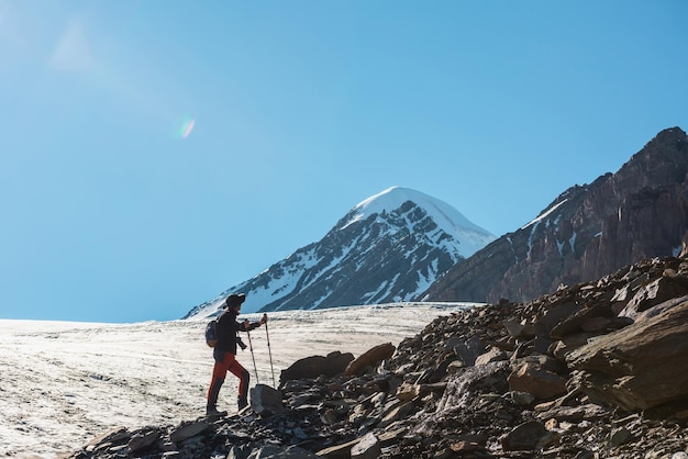 Paisagem alpina cênica com silhueta de caminhante com bastões de trekking contra grande língua de geleira e pico de montanha de neve à luz do sol Homem com mochila em altas montanhas sob o céu azul em dia ensolarado