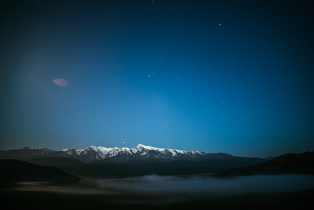 Paisagem atmosférica noturna com nevoeiro denso e grande cume da montanha nevada sob o céu estrelado. cenário alpino com nevoeiro espesso e grande cordilheira na noite estrelada. auge da neve sob o céu noturno de estrelas.