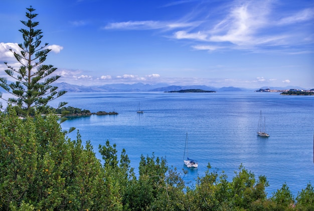 Paisagem bonita do verão - superfície calma da água do mar, iates brancos, árvores verdes e montanhas no horizonte. Ilha de Corfu, Grécia.