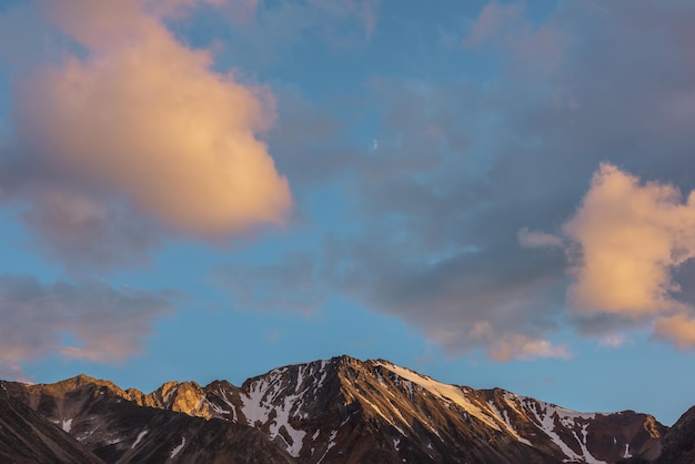 Paisagem cênica com alta montanha de neve com pináculo rochoso afiado na luz do sol dourada sob nuvens de cor do sol em clima mutável Vista colorida para o topo da montanha grande sob nuvens laranja no céu