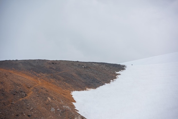 Paisagem cênica com trilha de caminhada na encosta de pedra para alta montanha de neve na luz do sol sob céu nublado cinza Caminho turístico na encosta rochosa iluminada pelo sol para grande montanha de neve em clima mutável