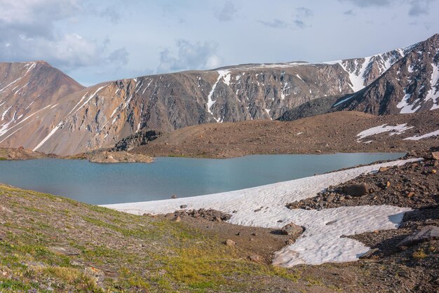 Paisagem colorida com belo lago no vale da montanha iluminada pelo sol perto da alta cordilheira sob céu nublado em clima variável Cenário impressionante com lago alpino entre rochas e neve na luz solar