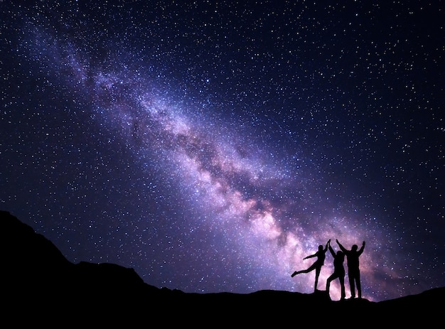 Paisagem com a Via Láctea roxa. Céu estrelado à noite com a silhueta de uma família feliz com os braços erguidos na montanha.