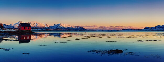 Paisagem com bela casa de rorbu vermelho do lago de inverno e montanhas nevadas ao pôr do sol nas Ilhas Lofoten no norte da Noruega Vista panorâmica