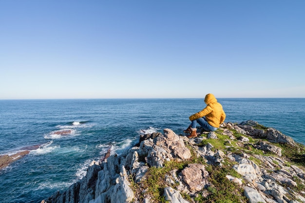 Paisagem costeira de rochas e mar com mulher sentada olhando para o mar com jaqueta amarela com capuz Copiar espaço