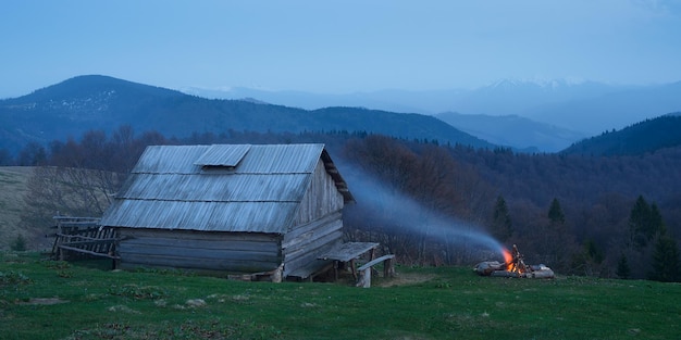 Paisagem da noite com casa de madeira e o fogo. Acampar nas montanhas. Crepúsculo após o pôr do sol. Montanhas dos Cárpatos, Ucrânia, Europa