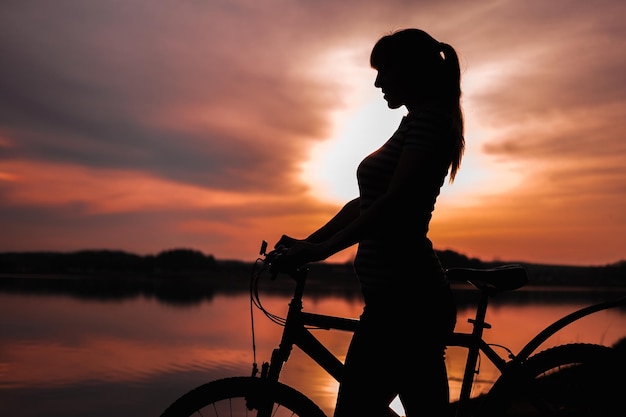 Paisagem de silhueta de verão Garota com uma bicicleta à beira do lago contra o pano de fundo do sol poente o pôr do sol carmesim