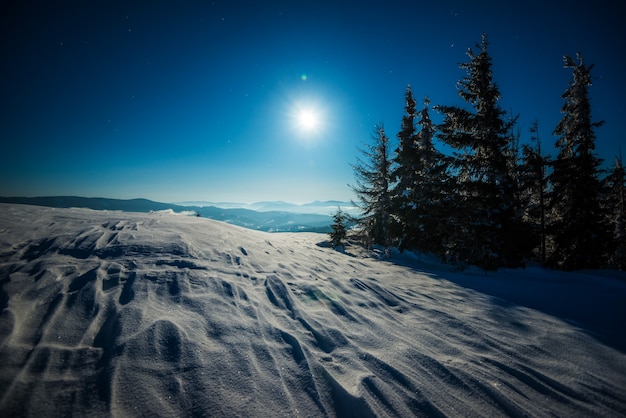 Paisagem de uma pista de esqui com neve ao fundo da floresta de abetos e cadeias de montanhas ao luar