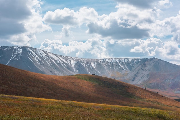 Paisagem heterogênea de outono com colinas em planaltos altos e cordilheira nevada iluminada pelo sol sob céu nublado dramático Cores vívidas de outono nas montanhas Luz solar e sombras de nuvens em clima variável