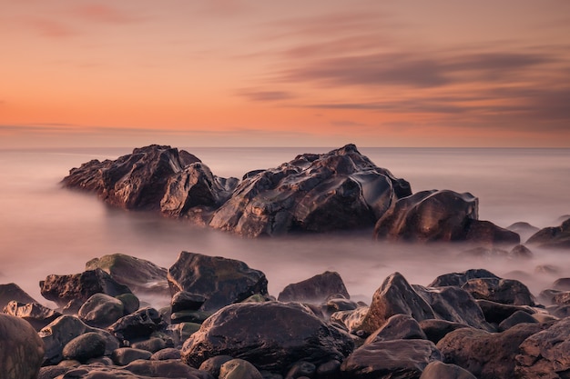 Paisagem marítima de longa exposição ao nascer do sol mostrando rochas perto da costa com o brilho do sol nas pedras úmidas e água turva leitosa
