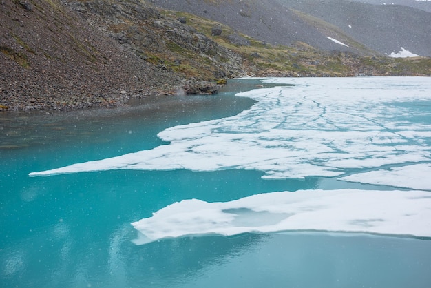 Paisagem montanhosa atmosférica com lago alpino congelado durante a queda de neve Gelo claro flutua na superfície da água do lago Cenário montanhoso impressionante com lago gelado da montanha Flocos de neve no fundo da água