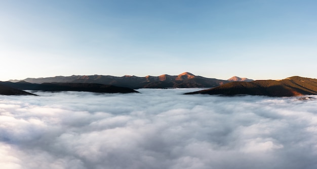 Paisagem montanhosa de nevoeiro acima das nuvens ao pôr do sol. Vista aérea. Os picos das montanhas sobressaem das nuvens.