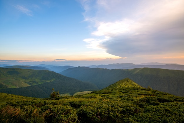 Paisagem montanhosa de noite de verão com colinas gramadas e picos distantes ao pôr do sol colorido.