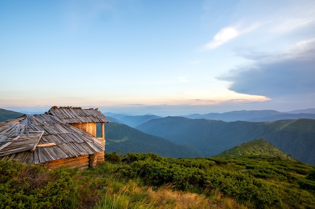 Paisagem montanhosa de noite de verão com o antigo abrigo turístico abandonado em colinas gramadas e picos distantes ao pôr do sol colorido.