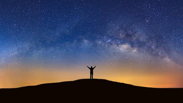 Paisagem panorâmica com via láctea Céu noturno com estrelas e silhueta de um homem desportivo em pé com braços levantados em alta montanha