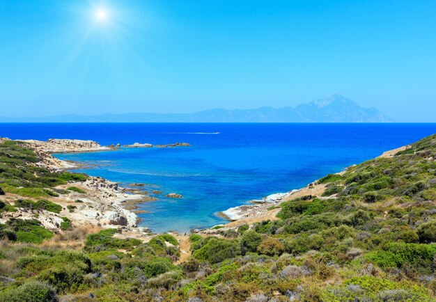 Paisagem pedregosa da costa do mar ensolarado do verão com vista de montagem de Athos em distante (Halkidiki, Sithonia, Grécia).