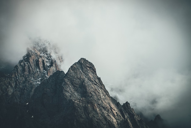 Paisagem surreal atmosférica escura com topo de montanha rochosa escura em nuvens baixas no céu nublado cinza. Nuvem baixa cinzenta no alto pináculo. Alta rocha negra com neve em nuvens baixas. Montanhas sombrias surrealistas.