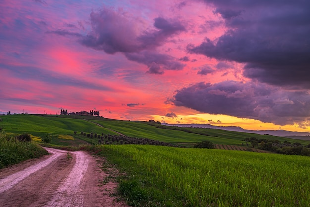 Paisagem verde única na Toscana, Itália. Céu dramático do pôr-do-sol, estrada de terra cruzando montanhas cultivadas e campos de cultivo de cereais.