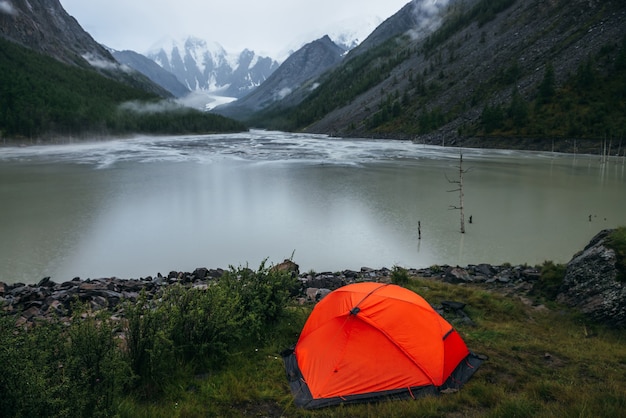 Paisaje alpino atmosférico con carpa naranja en la orilla del lago de montaña verde y montañas nevadas en tiempo lluvioso. Paisaje sombrío con círculos lluviosos sobre el agua del lago de montaña y nubes bajas en el valle