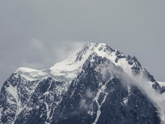 Paisaje alpino atmosférico con la cima de la montaña cubierta de nieve bajo el cielo gris nieve. Un paisaje impresionante con un hermoso pico puntiagudo con nieve y una pared de montaña nevada con nubes bajas.