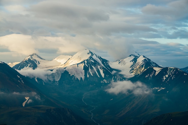 Paisaje alpino atmosférico con grandes montañas nevadas entre nubes bajas y valle de las tierras altas con corriente de agua de deshielo en hora dorada. Maravilloso paisaje al glaciar masivo en la cordillera gigante al amanecer