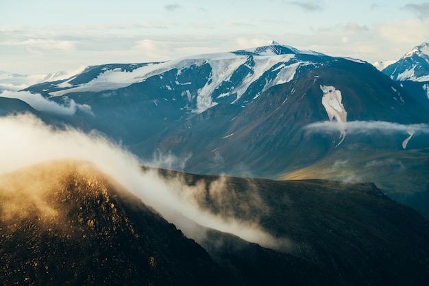 Paisaje alpino atmosférico con grandes nubes bajas doradas brillantes sobre roca y gigantes montañas nevadas con glaciares al amanecer. Volando sobre las montañas por encima de las nubes en la hora dorada. Maravilloso paisaje de montaña.