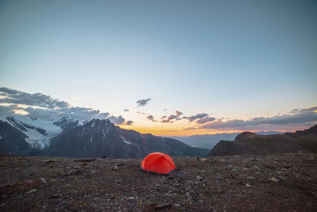 Paisaje alpino escénico con tienda de campaña a una altitud muy alta con vistas a grandes montañas en el cielo naranja del amanecer tienda de campaña naranja vívida con impresionante vista a la alta cordillera bajo un cielo nublado en los colores del atardecer