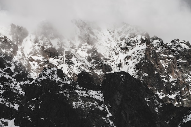 Paisaje alpino minimalista atmosférico con pico nevado de las montañas rocosas. Nubes bajas cerca del rango nevado. Rocas con nieve en la niebla. Montañas escarpadas en la niebla. Majestuoso paisaje brumoso y brumoso a gran altitud