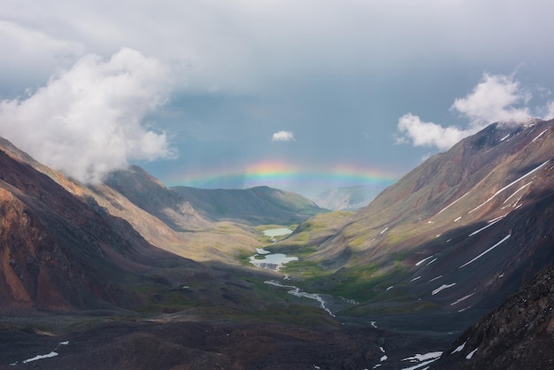 Paisaje alpino sombrío con arco iris vívido sobre el lago de montaña en el valle Paisaje sombrío con arco iris brillante sobre el lago en el valle de montaña Vista superior al arco iris colorido y nubes bajas en las montañas