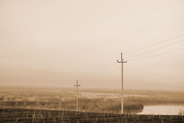 Paisaje atmosférico con líneas eléctricas en campo en el fondo del río bajo el cielo sepia.