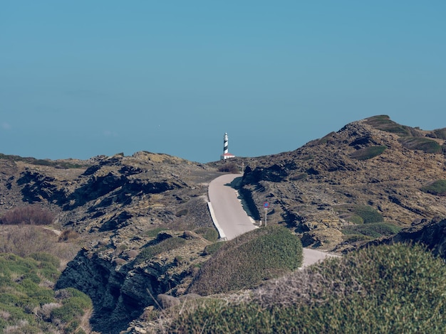 Paisaje de carretera entre tierras altas rocosas y hierba verde en colinas y faro en la cima bajo un cielo azul