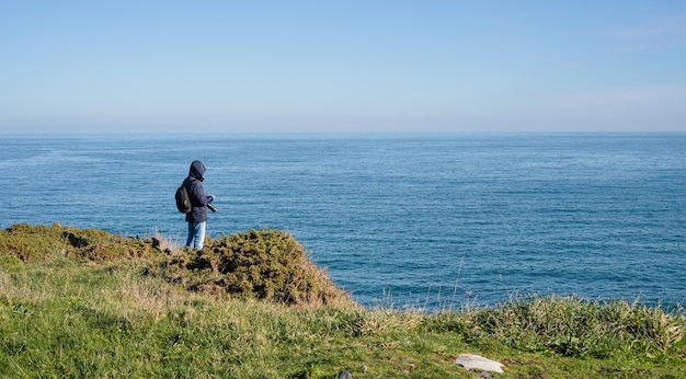 Paisaje costero y hombre con abrigo oscuro con capucha y cámara mirando el mar en calma Copiar espacio