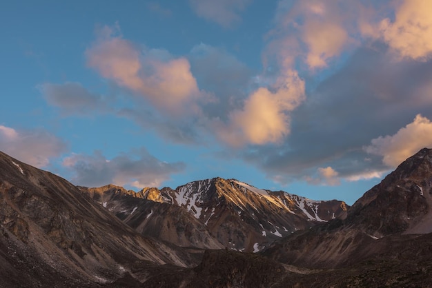 Paisaje escénico con alta montaña nevada con un pináculo rocoso afilado a la luz del sol dorada bajo nubes de color puesta de sol en un clima cambiante Vista colorida a la cima de la gran montaña bajo nubes naranjas en el cielo